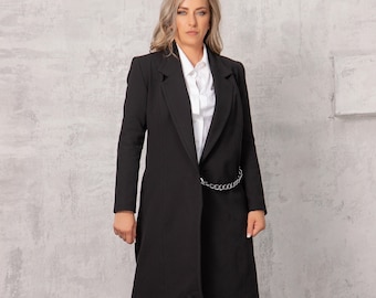 May include: A woman wearing a long black blazer with a silver chain belt, light blue jeans, and black boots. She is standing in front of a grey concrete wall.