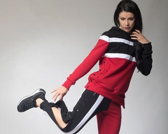 Peut inclure: Une femme portant un sweat à capuche rayé rouge, noir et blanc et un pantalon de survêtement assorti. Le pantalon de survêtement a une bande blanche sur le côté et le mot "A" sur la jambe. Elle porte des baskets noires.