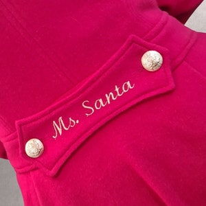 Op de afbeelding: Een rode jas met een gouden knoop en geborduurde tekst die "Ms. Santa" op de rug zegt.