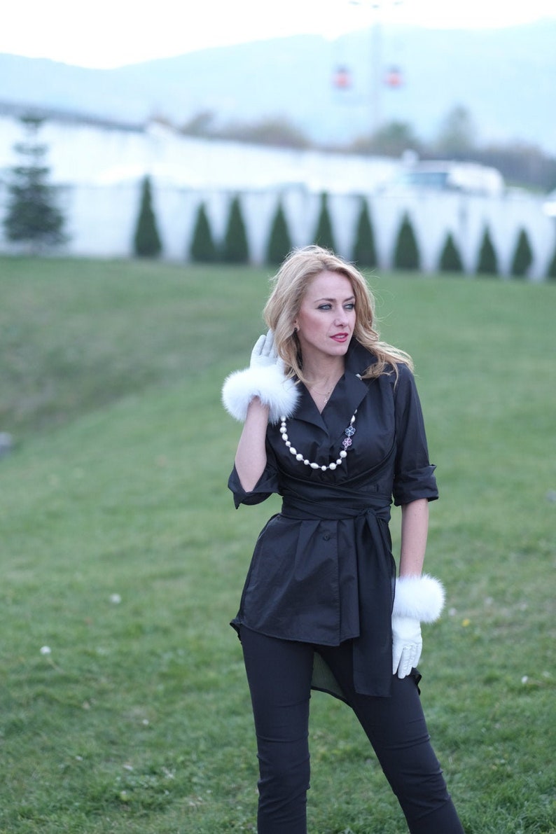 May include: A woman wearing a black button-down shirt with a pearl necklace and white fur gloves. She is standing in a grassy field.