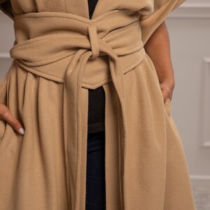 May include: A long, tan coat with a self-tie belt. The coat has a V-neck and long sleeves. The belt is tied in a knot at the front of the coat.