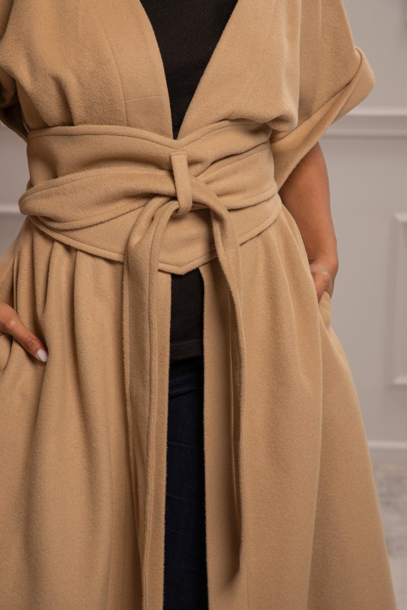 May include: A long, tan coat with a self-tie belt. The coat has a V-neck and long sleeves. The belt is tied in a knot at the front of the coat.