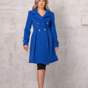 May include: A woman wearing a royal blue double-breasted coat with silver buttons. She is standing in front of a gray textured wall.