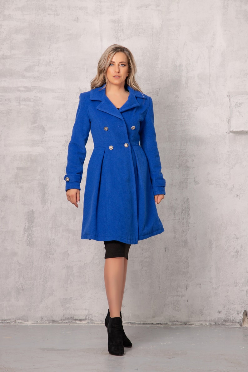 May include: A woman wearing a royal blue double-breasted coat with silver buttons. She is standing in front of a gray textured wall.