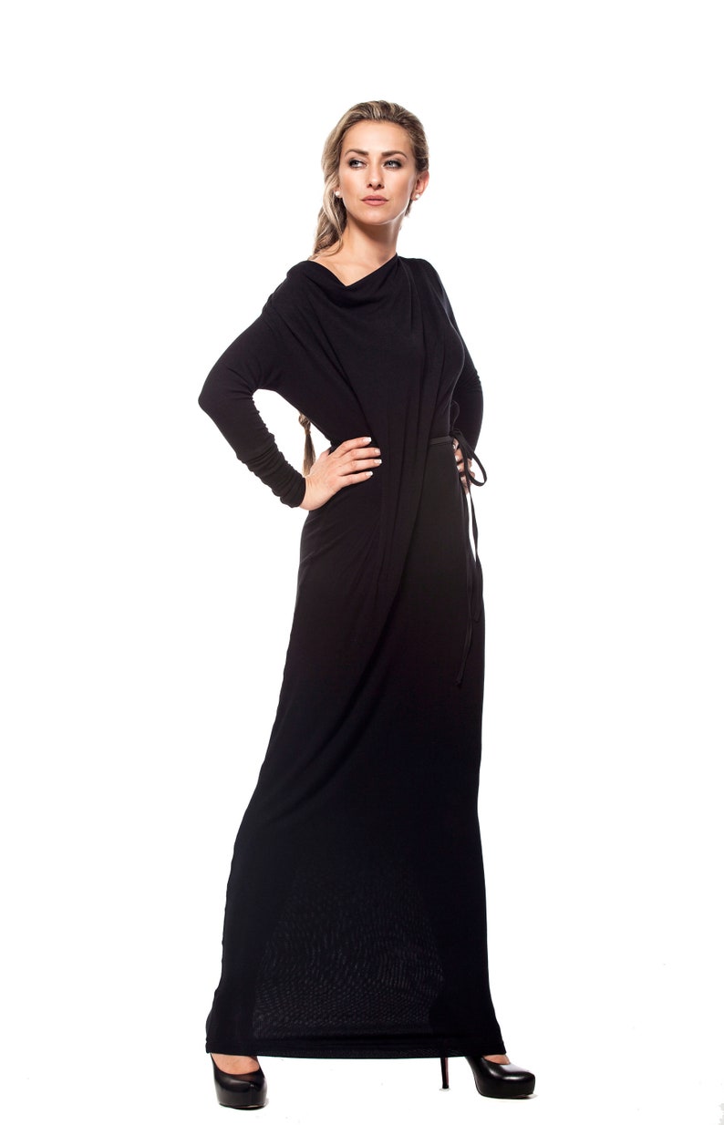 May include: A woman wearing a long black dress with a cowl neck and a belt. She is standing with her hand on her hip and looking to the side.