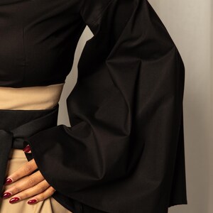 Op de afbeelding: Zwarte top met wijde, uitlopende mouwen. De outfit omvat een beige rok en een zwarte riem om de taille. De mouwen, met hun dramatische vorm, trekken de aandacht.