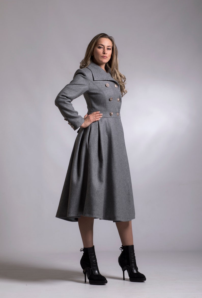 May include: A gray double-breasted coat with gold buttons and a flared skirt. The coat is worn by a person with long brown hair and is standing on a gray background.