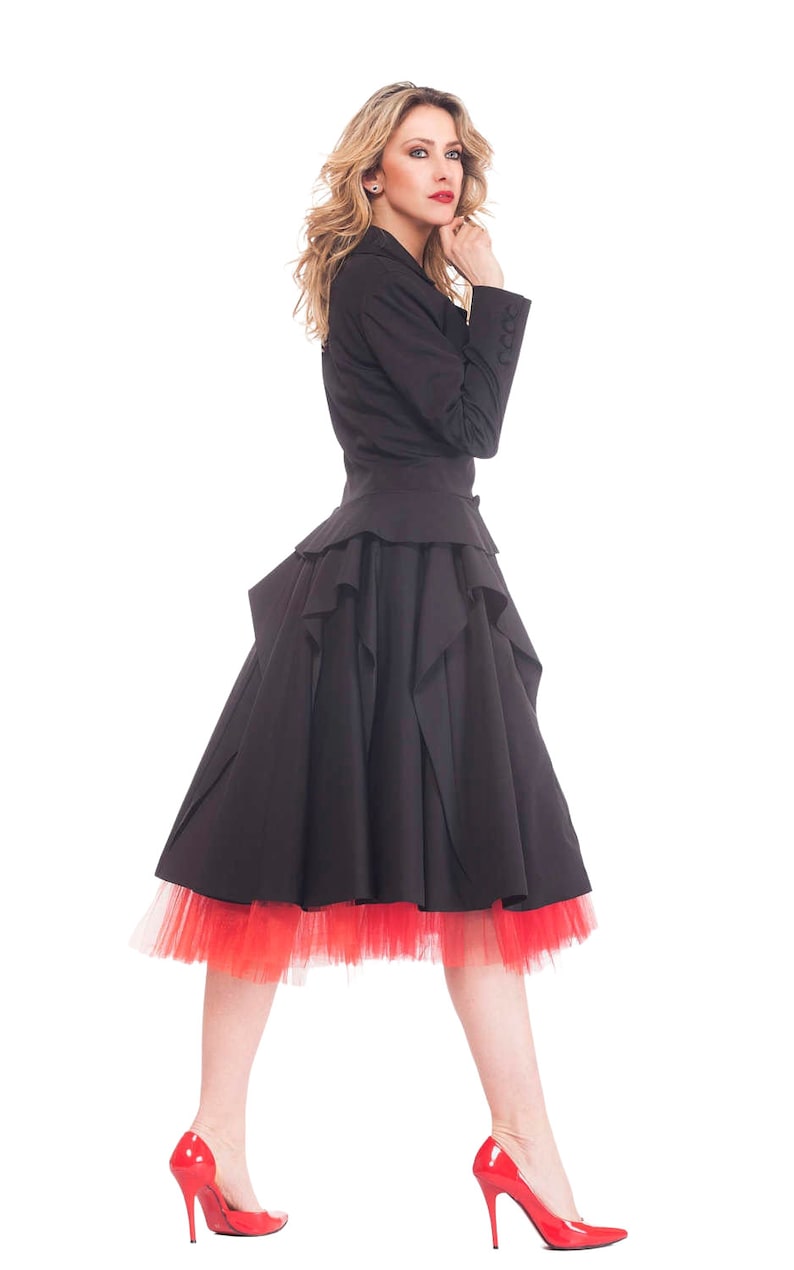 May include: A woman wearing a black dress with a ruffled skirt and a red tulle underskirt. She is wearing red high heels and has her hair styled in loose waves.