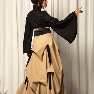 Op de afbeelding: Een zwarte top met wijde mouwen en een hoge kraag, gecombineerd met een beige rok met unieke draperie&euml;n en zwarte accenten. De outfit wordt gecompleteerd met een zwarte riem en hoge hakken.