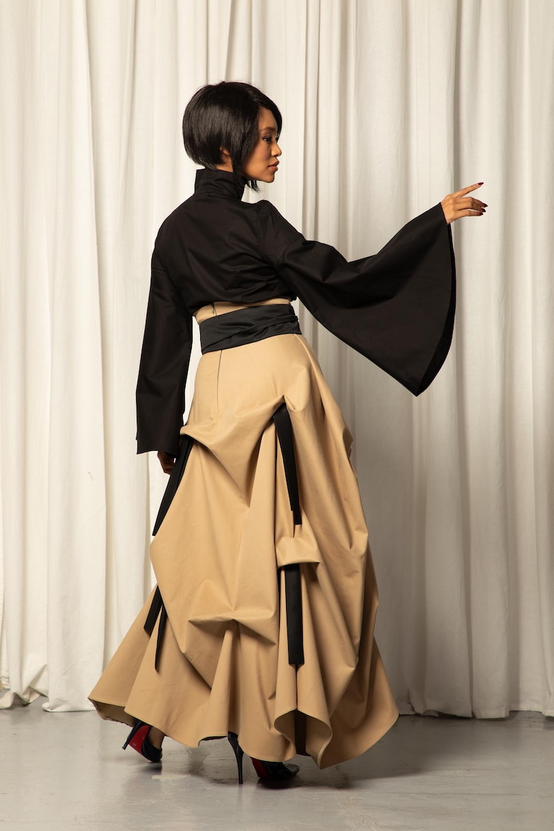 Op de afbeelding: Een zwarte top met wijde mouwen en een hoge kraag, gecombineerd met een beige rok met unieke draperie&euml;n en zwarte accenten. De outfit wordt gecompleteerd met een zwarte riem en hoge hakken.