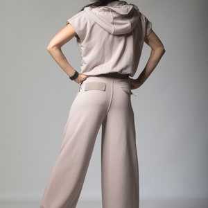 Puede incluir: Una mujer lleva una sudadera con capucha de color rosa claro con mangas cortas y pantalones anchos a juego. Los pantalones tienen un bolsillo de parche en la parte posterior.
