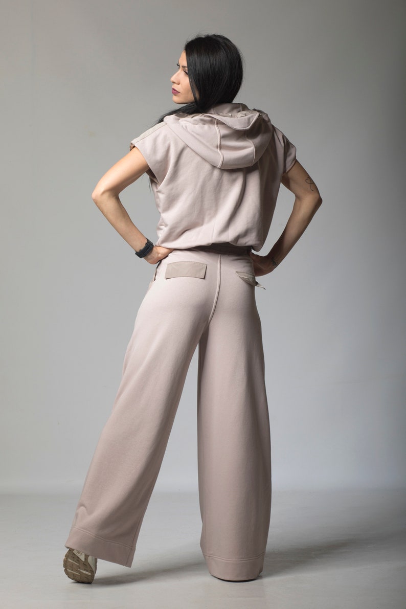 Puede incluir: Una mujer lleva una sudadera con capucha de color rosa claro con mangas cortas y pantalones anchos a juego. Los pantalones tienen un bolsillo de parche en la parte posterior.