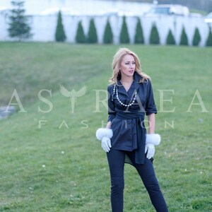 May include: A woman wearing a black wrap shirt with a pearl necklace and white fur gloves. She is standing on a sidewalk with green grass in the background.