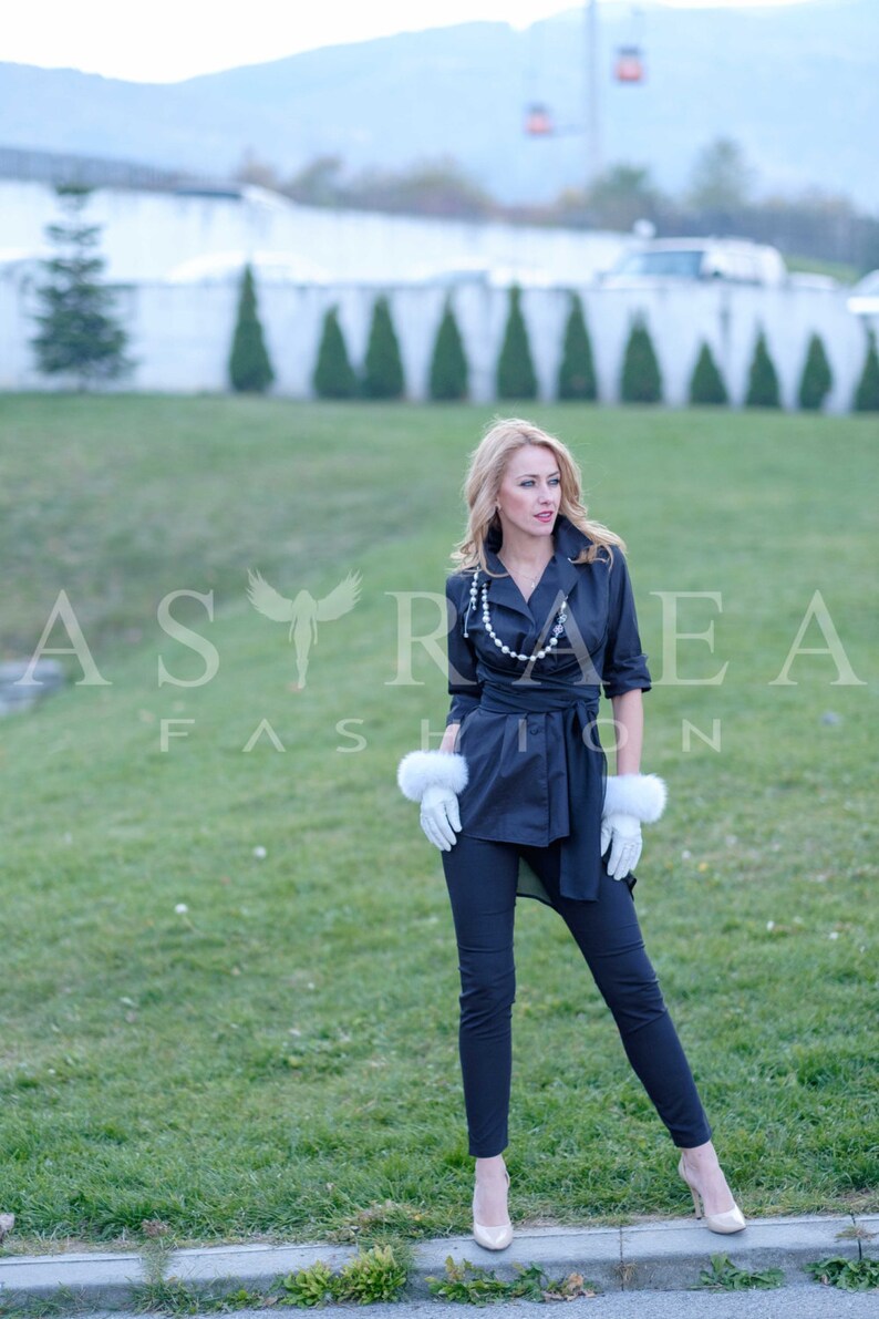 May include: A woman wearing a black wrap shirt with a pearl necklace and white fur gloves. She is standing on a sidewalk with green grass in the background.