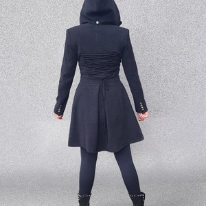May include: A dark gray hooded coat with a fitted waist and flared skirt. The back features a corset-style lace-up detail. The coat has long sleeves with buttoned cuffs and a small button at the hood. The model is wearing black leggings and black boots.
