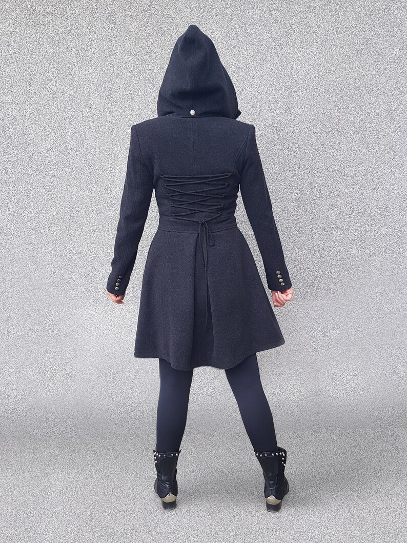May include: A dark gray hooded coat with a fitted waist and flared skirt. The back features a corset-style lace-up detail. The coat has long sleeves with buttoned cuffs and a small button at the hood. The model is wearing black leggings and black boots.