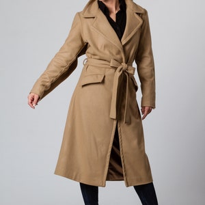 May include: A long camel-colored coat with a belt and a front pocket. The coat is worn by a person with dark hair and is standing on a light gray background.