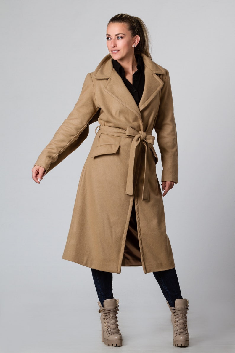 May include: A long camel-colored coat with a belt and a front pocket. The coat is worn by a person with dark hair and is standing on a light gray background.