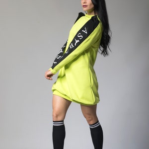 May include: A neon green sweatshirt dress with black sleeves and white lettering that reads "ASFR" on the left sleeve. The model is wearing black socks with white stripes and black sneakers.