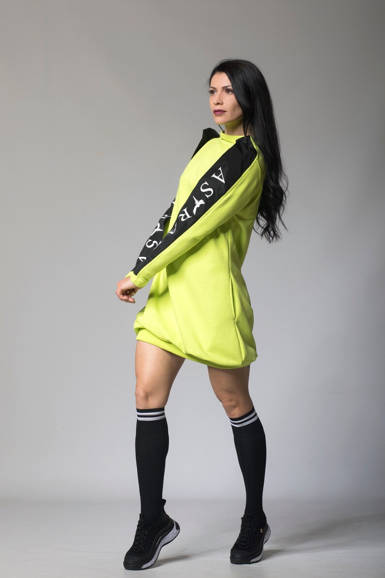 May include: A neon green sweatshirt dress with black sleeves and white lettering that reads "ASFR" on the left sleeve. The model is wearing black socks with white stripes and black sneakers.