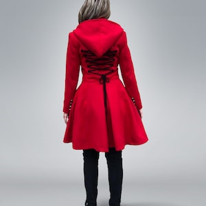 May include: A vibrant red hooded coat with a flared skirt, featuring a black lace-up back detail. The coat has long sleeves and is paired with black pants and sneakers. The overall design is a blend of classic and modern styles.