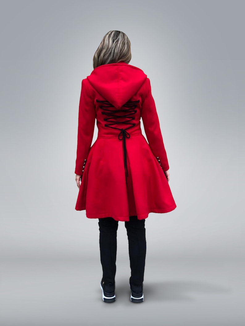 May include: A vibrant red hooded coat with a flared skirt, featuring a black lace-up back detail. The coat has long sleeves and is paired with black pants and sneakers. The overall design is a blend of classic and modern styles.