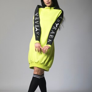 May include: A neon green sweatshirt dress with black sleeves featuring the text "ASRAEA" on each sleeve. The model is wearing black knee-high socks with white stripes and black sneakers.