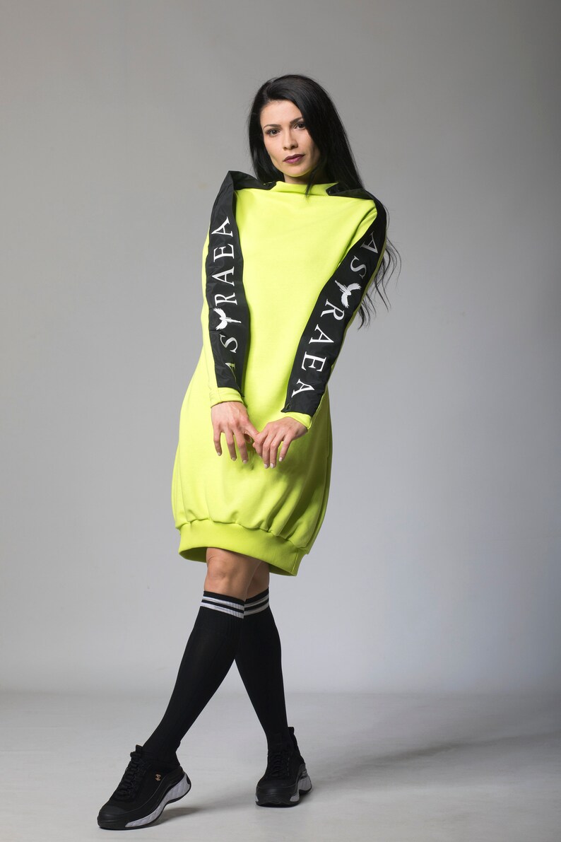 May include: A neon green sweatshirt dress with black sleeves featuring the text "ASRAEA" on each sleeve. The model is wearing black knee-high socks with white stripes and black sneakers.