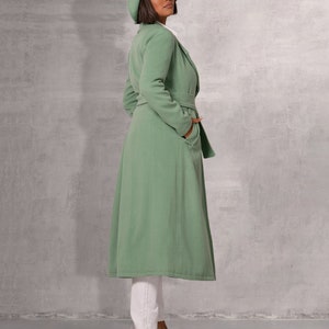 May include: A woman wearing a long, sage green coat with a matching hat. The coat has a belt and pockets. She is wearing white trousers and silver heels.