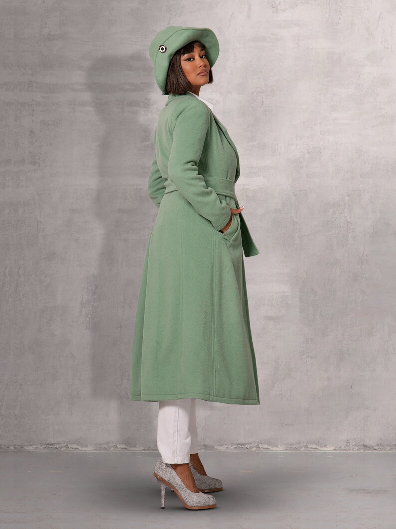 May include: A woman wearing a long, sage green coat with a matching hat. The coat has a belt and pockets. She is wearing white trousers and silver heels.