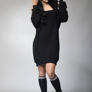 Puede incluir: Vestido negro con capucha y hombros descubiertos. El vestido tiene un cord&oacute;n y se combina con calcetines negros hasta la rodilla con rayas blancas y botas de plataforma negras. La persona lleva la capucha.