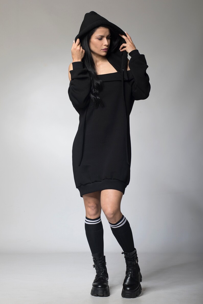 Puede incluir: Vestido negro con capucha y hombros descubiertos. El vestido tiene un cord&oacute;n y se combina con calcetines negros hasta la rodilla con rayas blancas y botas de plataforma negras. La persona lleva la capucha.