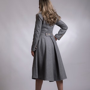 May include: A gray long-sleeved coat with a fitted bodice and a flared skirt. The coat has a high collar and a button closure. The model is wearing black heeled boots.