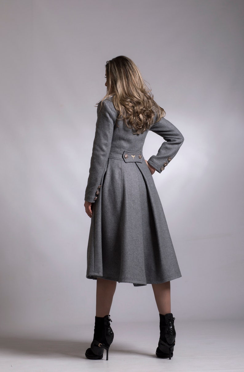 May include: A gray long-sleeved coat with a fitted bodice and a flared skirt. The coat has a high collar and a button closure. The model is wearing black heeled boots.