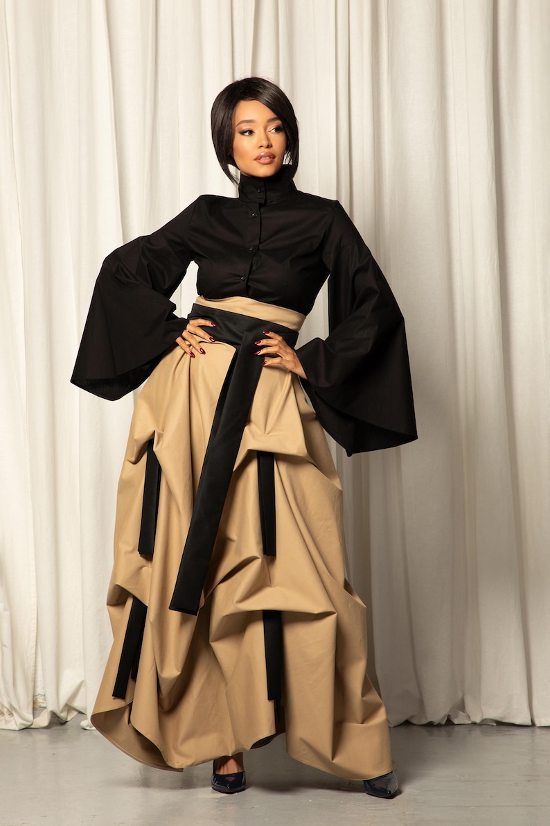 Op de afbeelding: Een zwarte top met hoge kraag en wijde mouwen, gecombineerd met een beige rok met zwarte details en een zwarte riem. De outfit is een moderne interpretatie van een klassieke stijl.