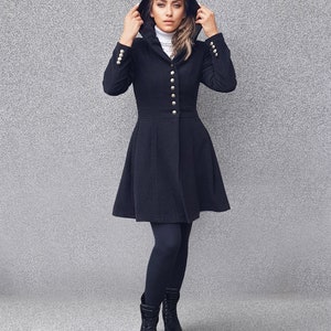 May include: A black hooded coat with gold buttons and a flared skirt. The coat is worn with black leggings and boots. The model is wearing a white turtleneck underneath the coat.