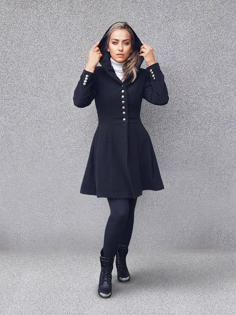 May include: A black hooded coat with gold buttons and a flared skirt. The coat is worn with black leggings and boots. The model is wearing a white turtleneck underneath the coat.