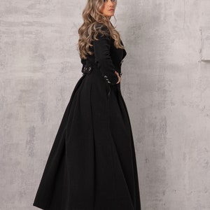 Pode incluir: Uma mulher vestindo um longo casaco preto com cinto e gola alta. O casaco tem uma saia fluida e &eacute; feito de um tecido macio e luxuoso.
