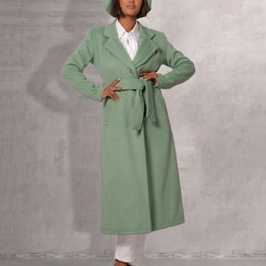 May include: A long, green coat with a belt and a button fastening. The coat has a collar and long sleeves. The model is wearing a white shirt and a green beret.