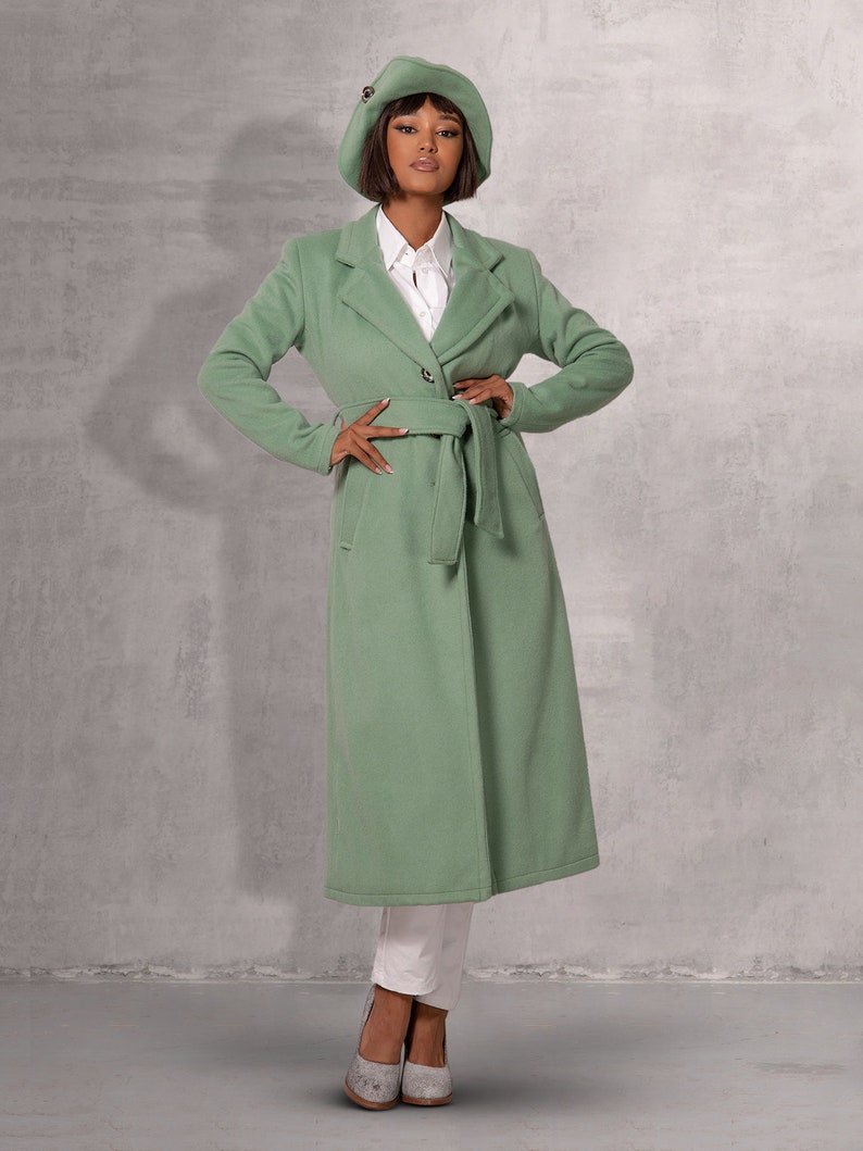 May include: A long, green coat with a belt and a button fastening. The coat has a collar and long sleeves. The model is wearing a white shirt and a green beret.
