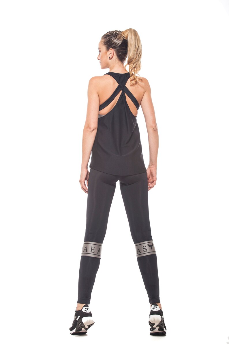 May include: Black tank top with a criss-cross back design and black leggings with silver lettering that says "AEA" on the left leg and "AS" on the right leg. The model is wearing black sneakers.