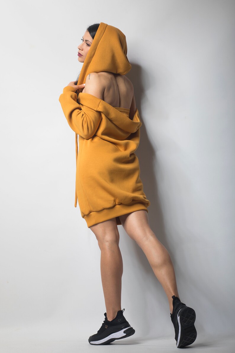 Puede incluir: Vestido con capucha de color amarillo mostaza. El vestido tiene un corte holgado, mangas largas y una capucha con cord&oacute;n. La modelo lleva zapatillas negras con suelas blancas. El fondo es una pared blanca lisa.