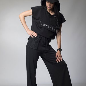 Puede incluir: Una sudadera con capucha negra de manga corta y cintura con cord&oacute;n. La sudadera tiene un logotipo blanco que dice "ASTRAEA" con un dise&ntilde;o de p&aacute;jaro estilizado. La modelo lleva pantalones negros de pierna ancha.