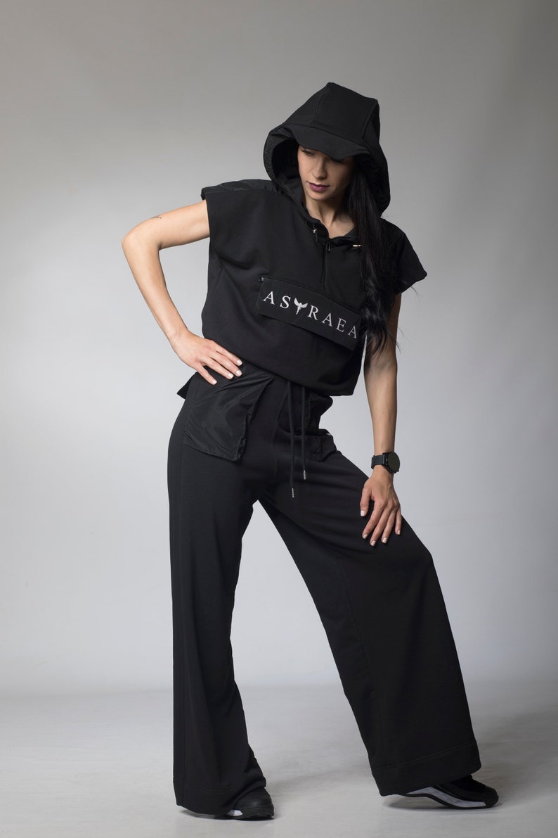 Puede incluir: Una sudadera con capucha negra de manga corta y cintura con cord&oacute;n. La sudadera tiene un logotipo blanco que dice "ASTRAEA" con un dise&ntilde;o de p&aacute;jaro estilizado. La modelo lleva pantalones negros de pierna ancha.