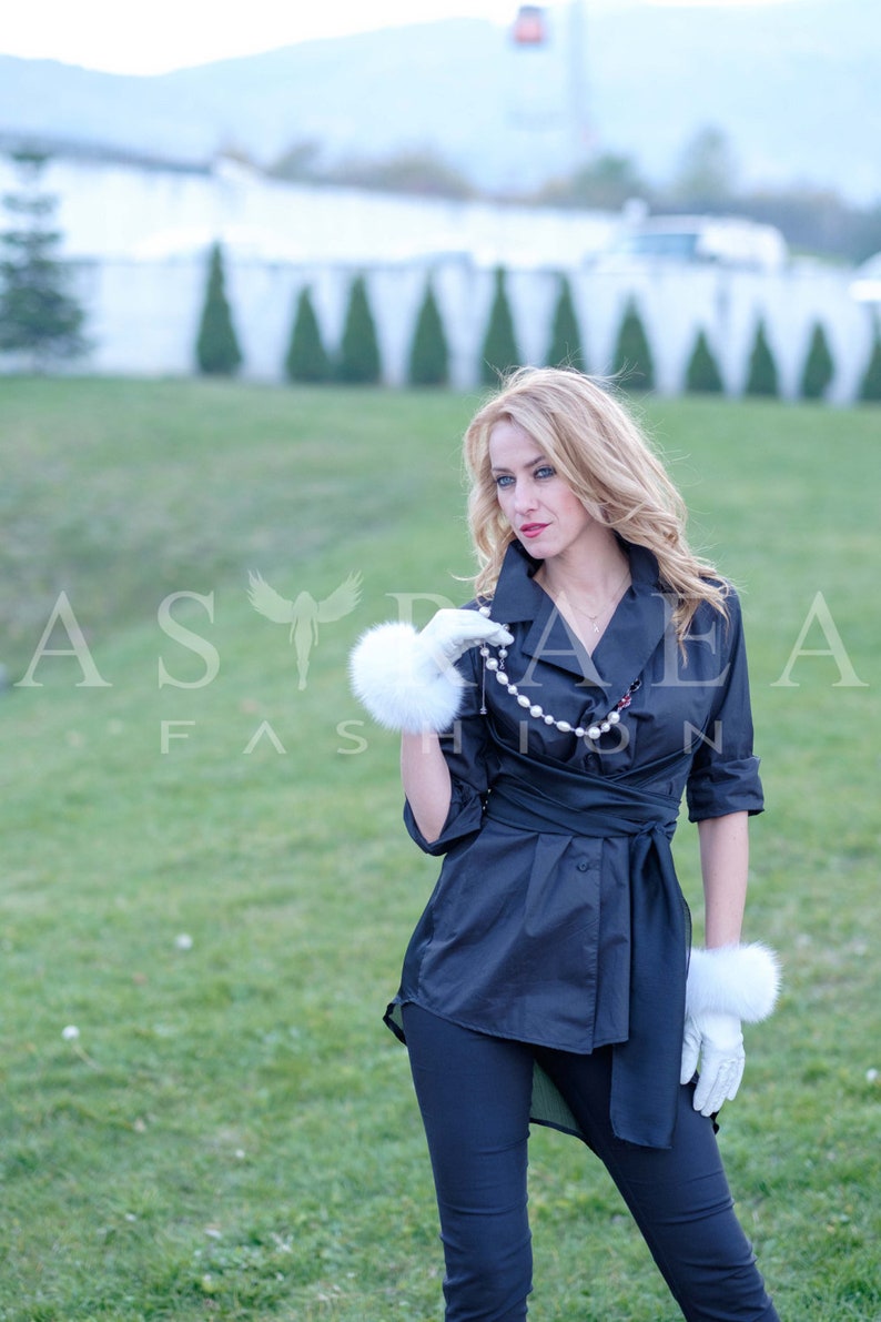May include: A woman wearing a black wrap shirt with a white pearl necklace and white fur gloves. She is standing in a grassy field.