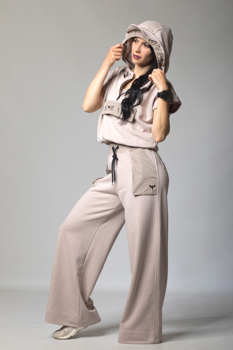 Puede incluir: Una sudadera con capucha beige claro con cremallera y un logotipo en el pecho, combinada con pantalones anchos a juego. Los pantalones tienen un bolsillo de parche grande en el lado derecho.