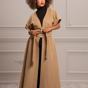 May include: A long, tan coat with a V-neck and a self-tie belt. The coat has short sleeves and a flowing skirt.