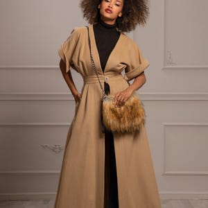 May include: A woman wearing a long, camel-colored coat with a fur-trimmed bag slung over her shoulder. She is wearing a black turtleneck sweater and dark blue jeans. The coat has a V-neckline and a single button closure at the waist. The woman is standing in front of a white wall.