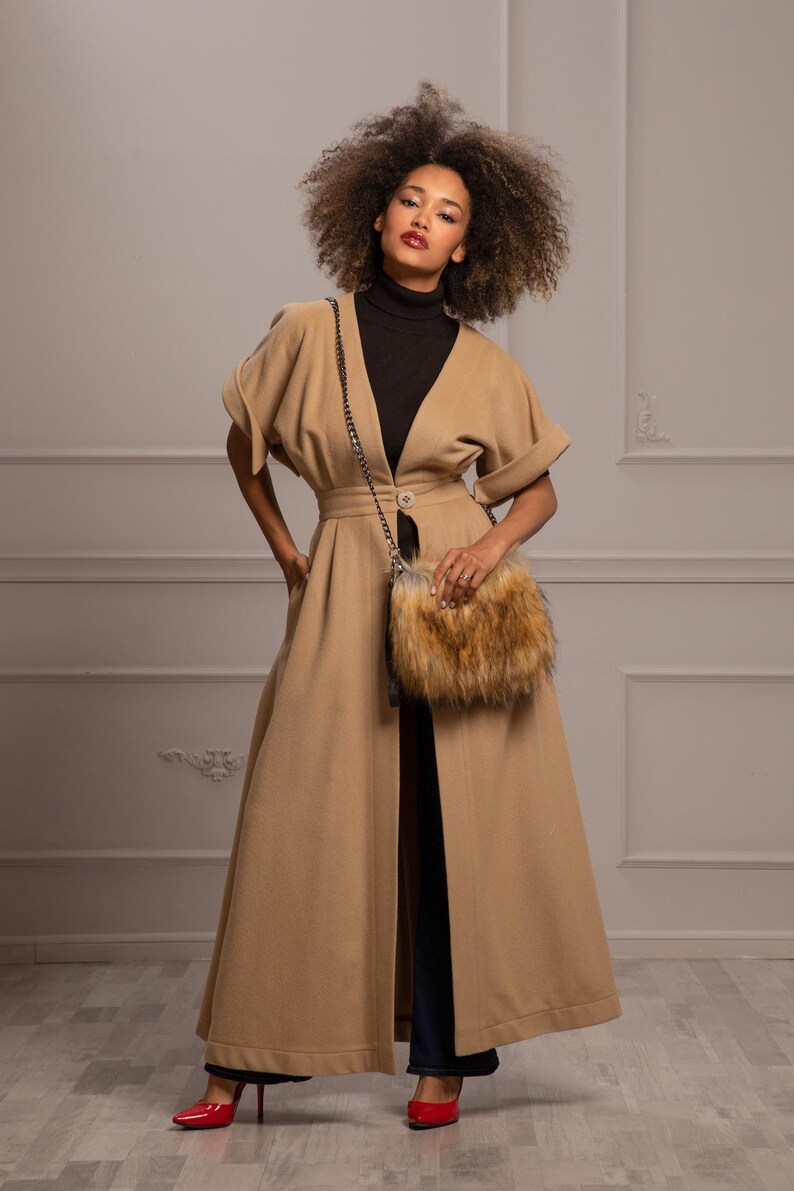 May include: A woman wearing a long, camel-colored coat with a fur-trimmed bag slung over her shoulder. She is wearing a black turtleneck sweater and dark blue jeans. The coat has a V-neckline and a single button closure at the waist. The woman is standing in front of a white wall.