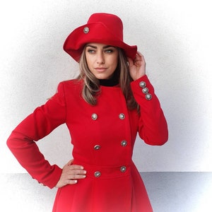 Puede incluir: Un abrigo rojo de doble botonadura con botones plateados y un sombrero rojo a juego. El abrigo tiene una falda acampanada y un cuello alto.